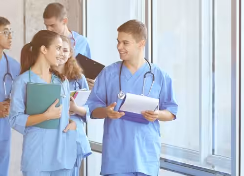 A group of five medical students in blue scrubs and stethoscopes walk and talk in a hallway.
