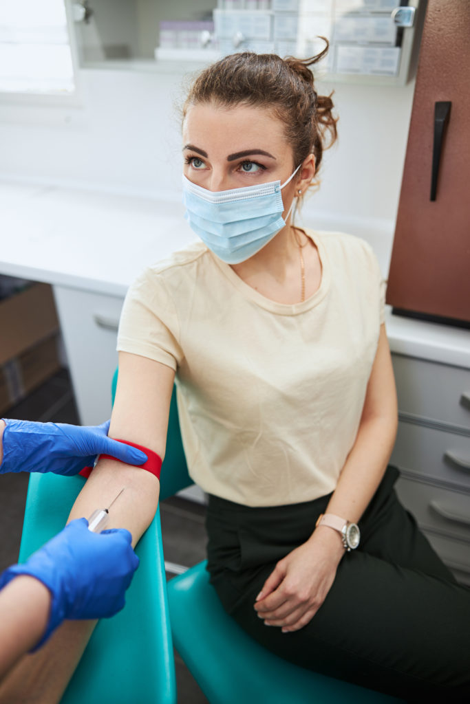Young female patient in a face mask sitting in the chair during the venipuncture performed by a nurse