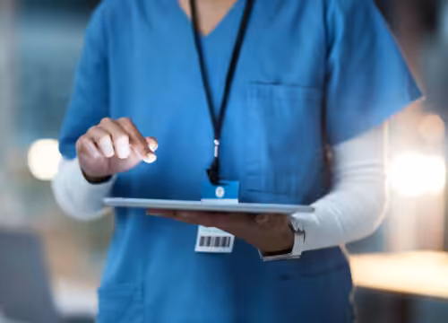 A person in blue scrubs holds and taps a silver tablet.
