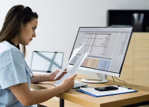 A person in light blue scrubs and a stethoscope looks at papers by a computer.