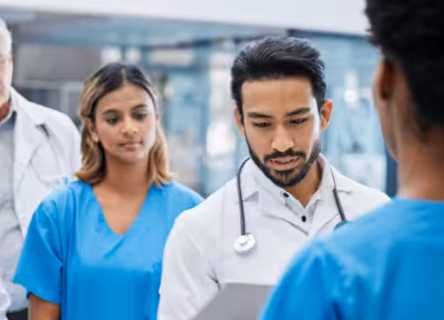 A group of medical professionals in scrubs and lab coats looks towards a person speaking.