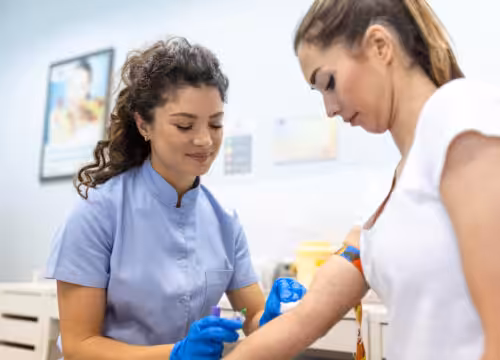 Phlebotomist in blue uniform and gloves draws blood from a patient's arm.