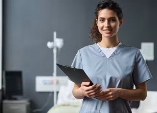 Smiling healthcare worker in gray scrubs holding a clipboard in a clinic exam room.