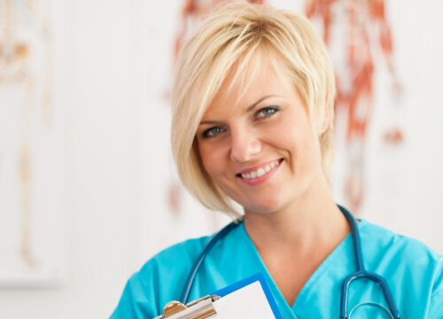 Blonde healthcare worker in blue scrubs smiles while holding a clipboard and wearing a stethoscope.