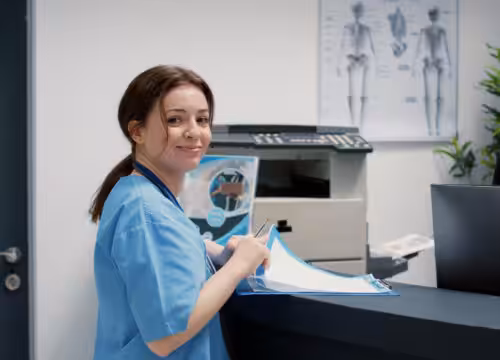 Medical assistant in blue scrubs holding patient forms at a clinic reception desk.